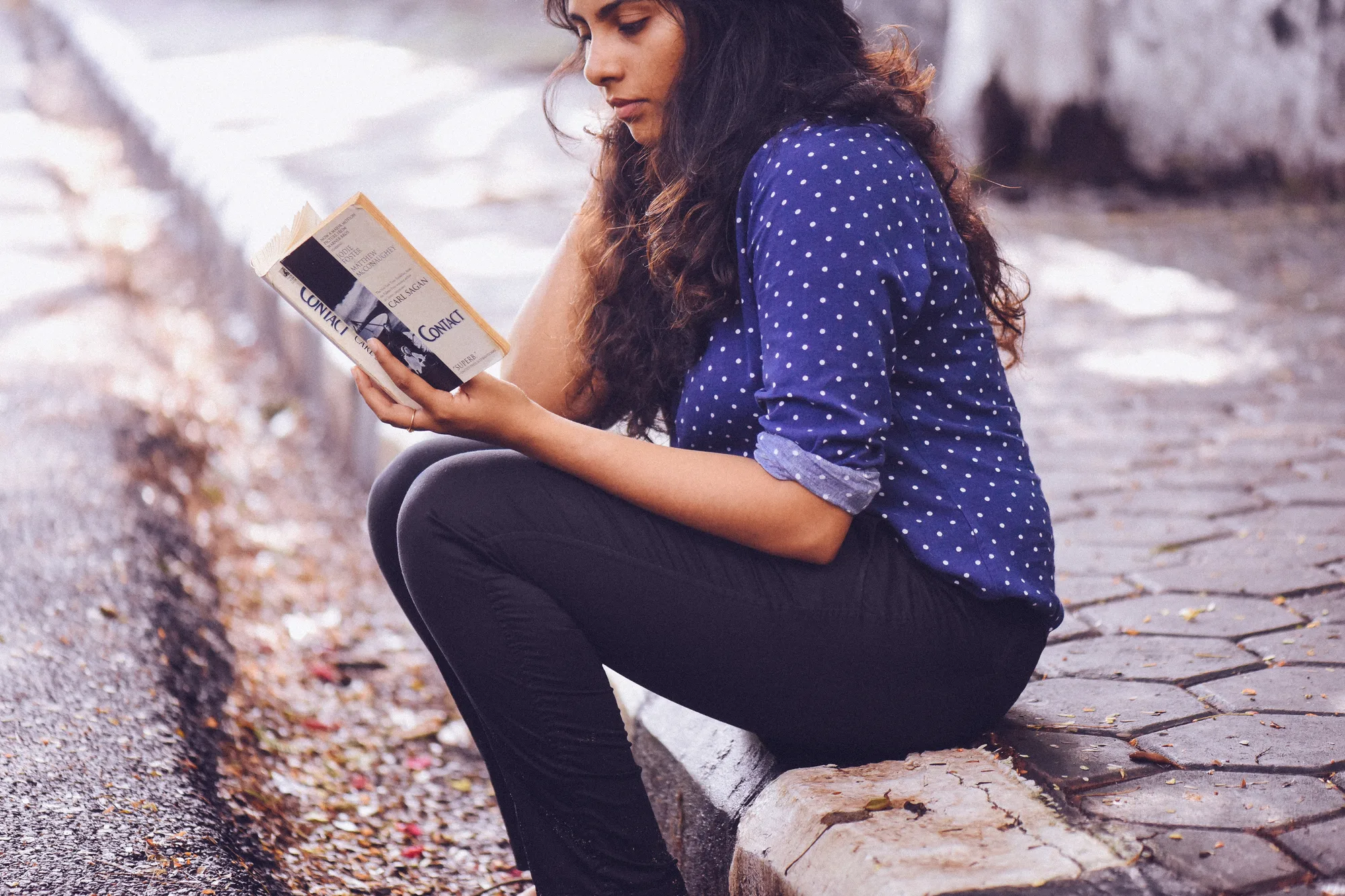 A young woman reading a book on a kerb.